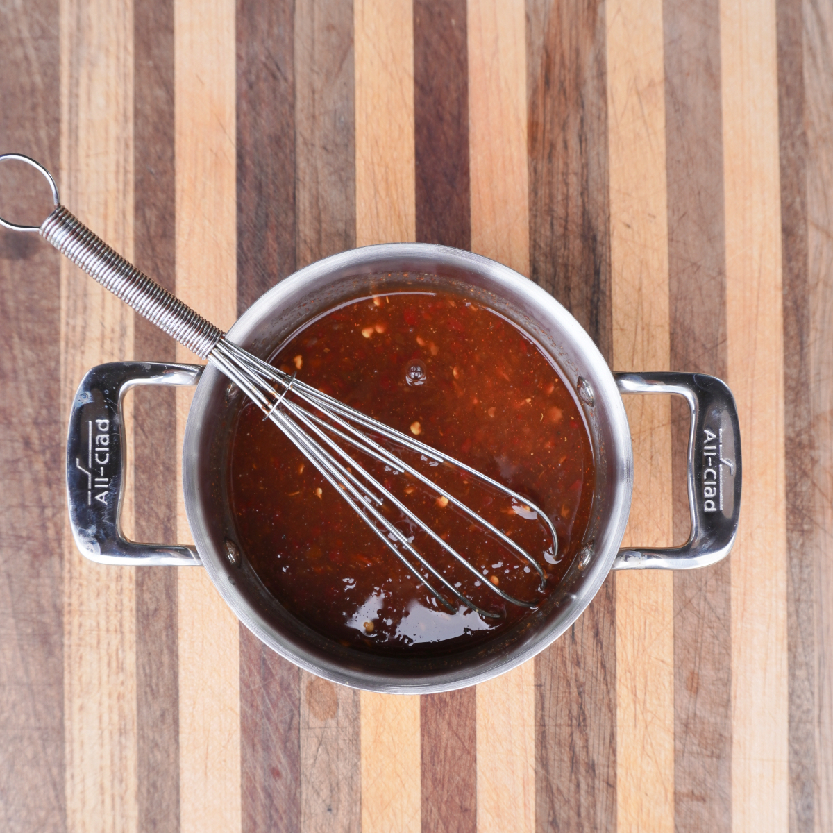 A sauce pan full of the sweet chili sauce and a whisk inside on top of a wooden cutting board.