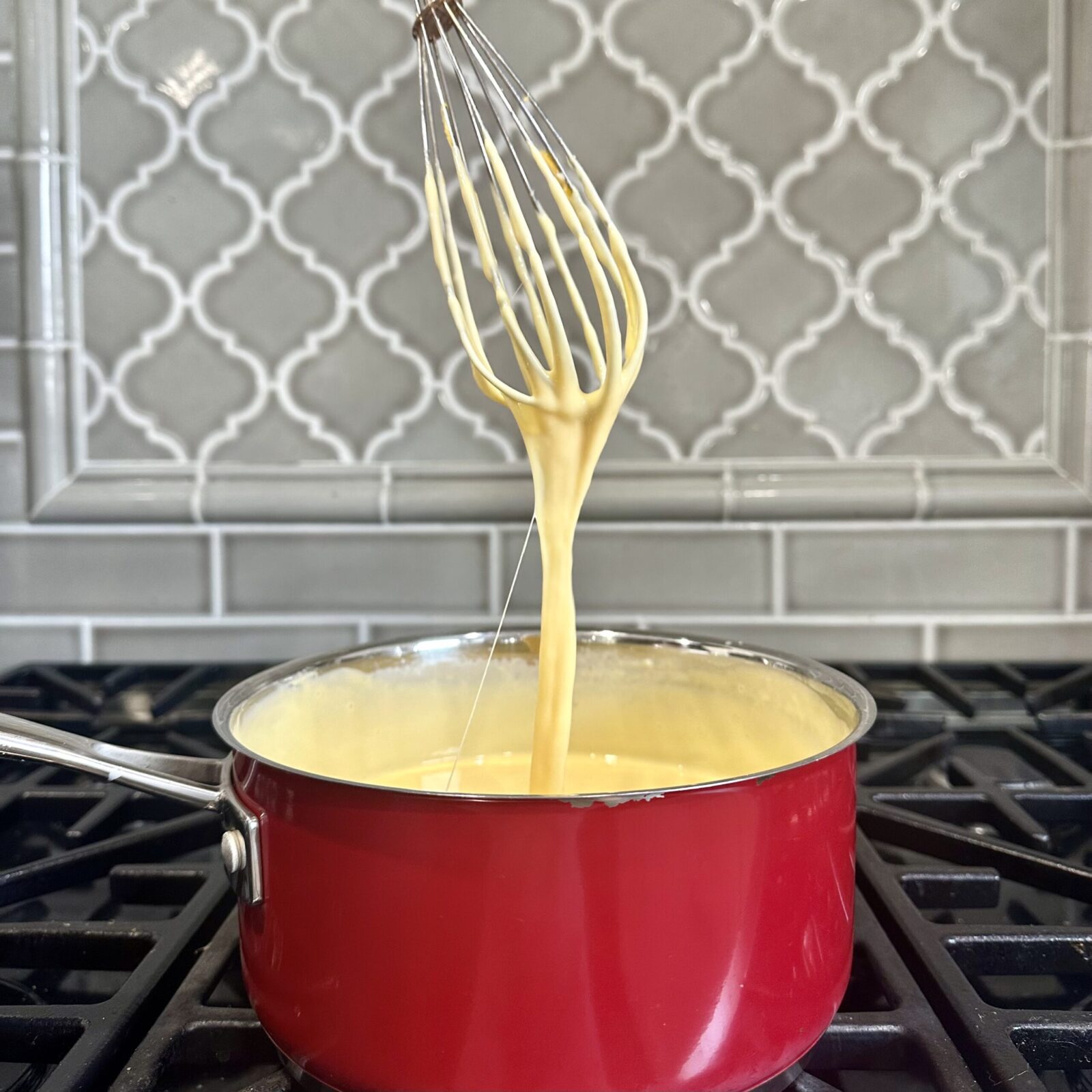 Creamy mac and cheese sauce stretched on a whisk from the sauce pan over the stove.