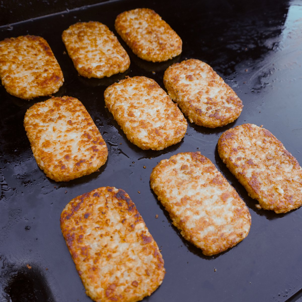 Three rows of hash browns grilled on a greasy griddle.
