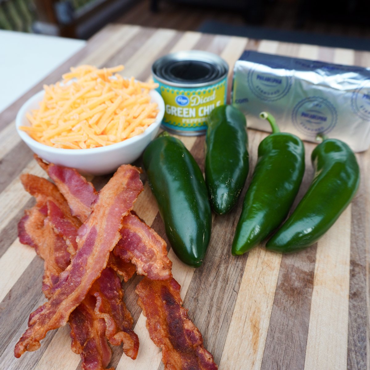 Jalapeno popper dip ingredients including jalapeno peppers, cooked bacon, shredded cheddar cheese, canned green chilies and a block of cream cheese.