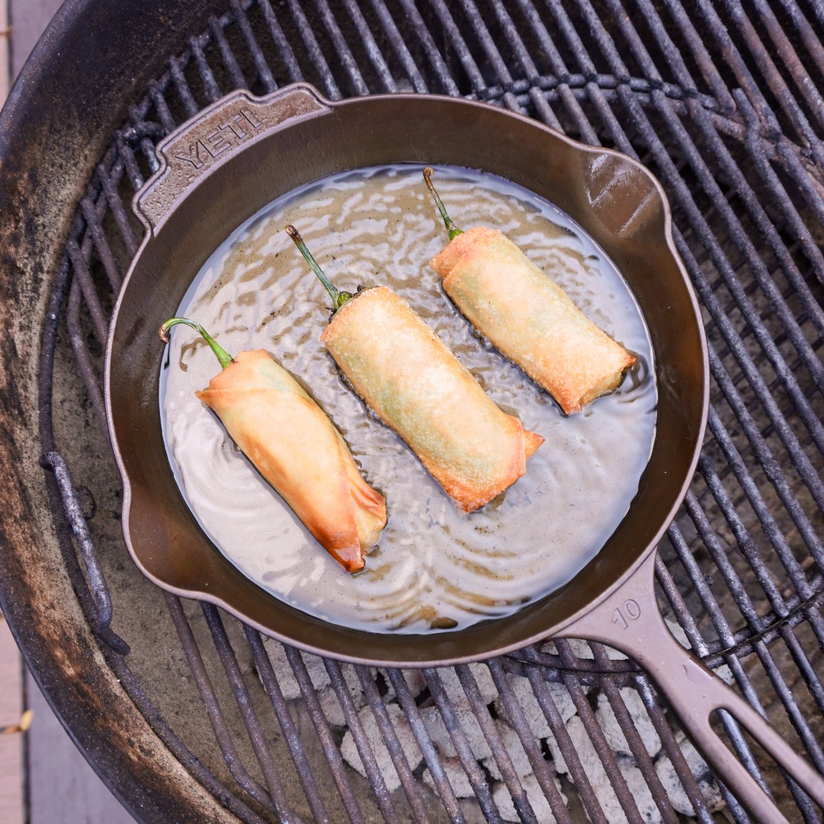 deep frying jalapeno popper lumpia's in a cast iron skillet on a charcoal grill.