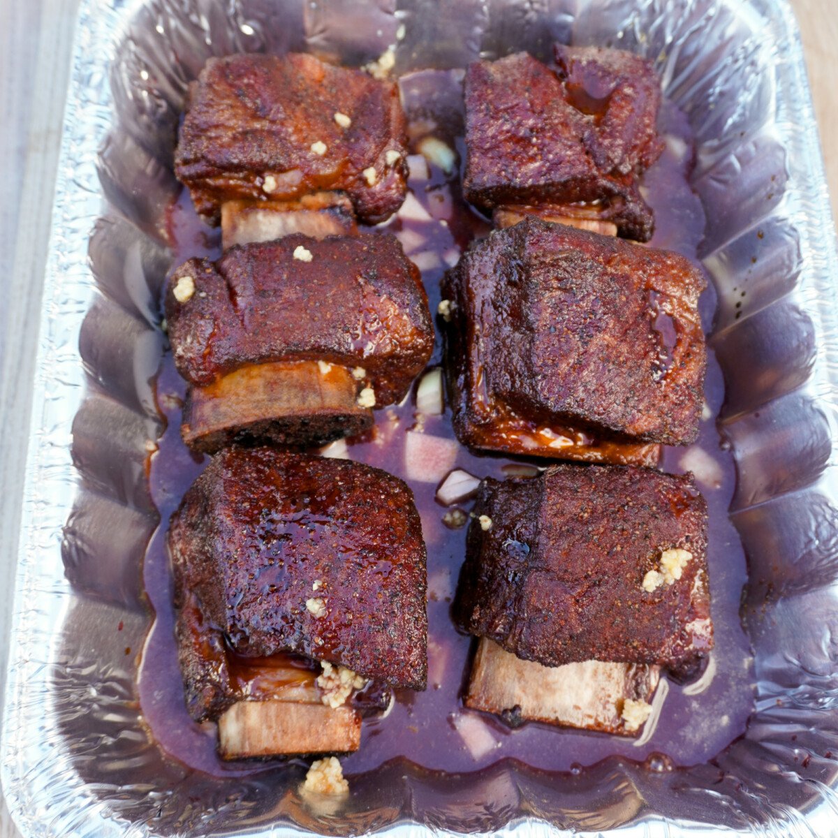 beef short ribs in a foil pan on a bed of onions with beef stock and red wine.