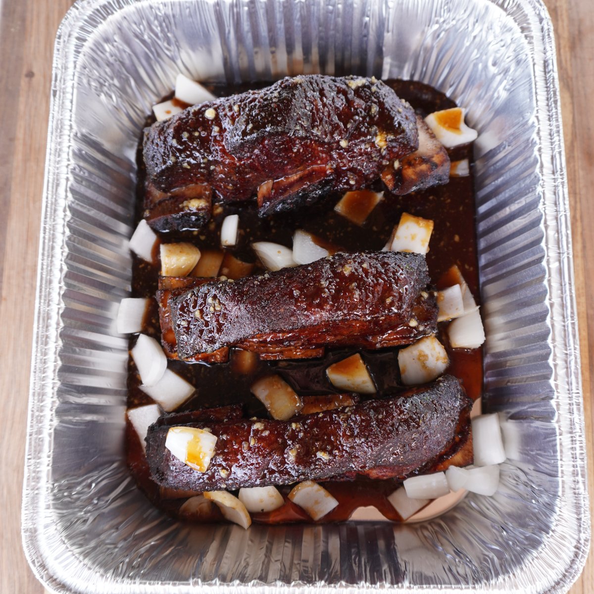 grilled beef short rib bites braised in liquid and onion in an aluminum pan.