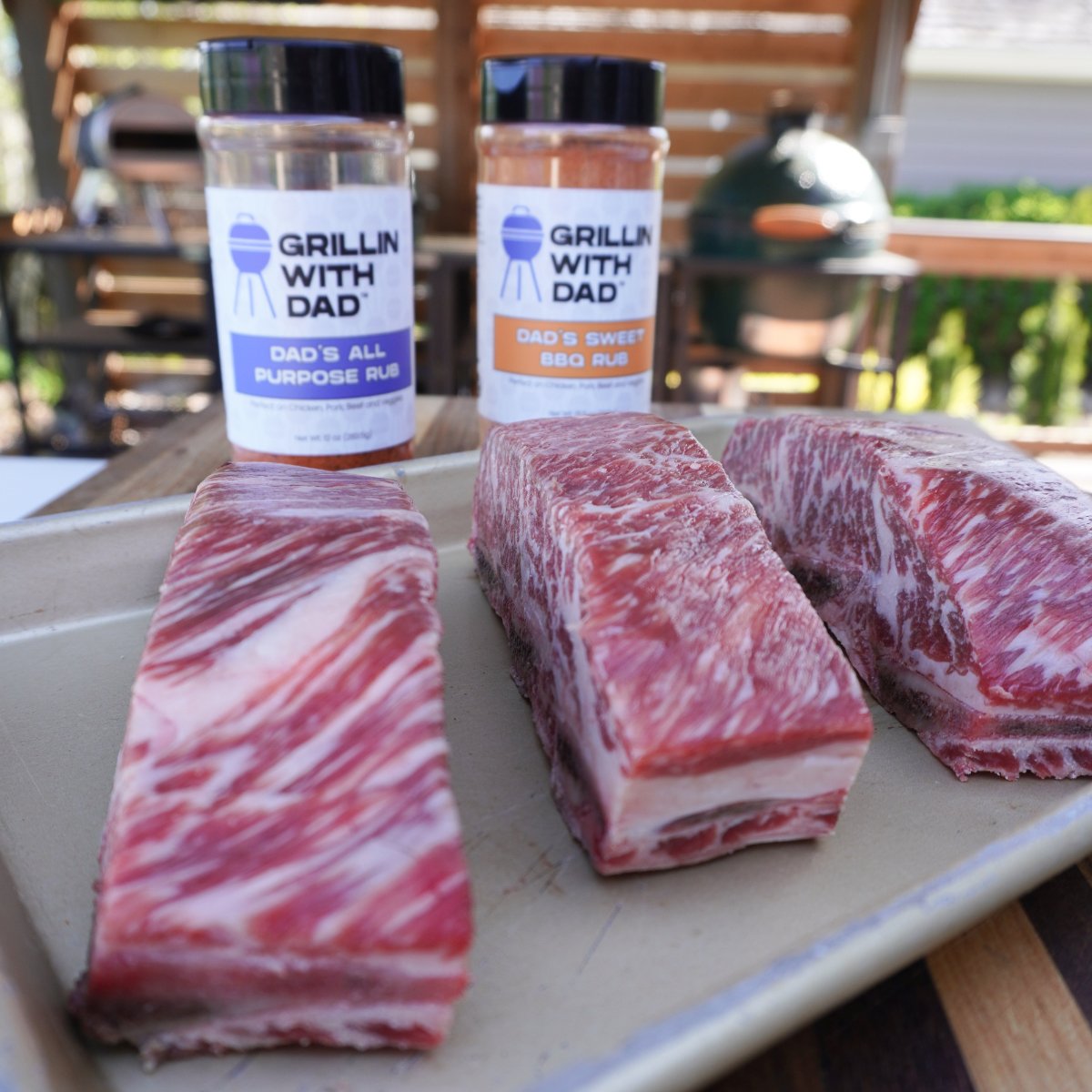 3 beef short ribs on a metal tray with Grillin with dad All Purpose rub and Sweet BBQ rub bottles standing behind the.